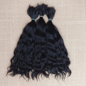May include: Three bundles of wavy, black hair tied with green rubber bands. The hair has a natural wave pattern and a dark, rich color, suggesting a hair extension or wig product. The background is a neutral, textured fabric.