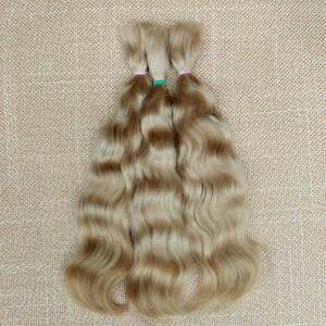 May include: Three bundles of wavy, light blonde hair with darker brown lowlights. Each bundle is tied with a small elastic band in pink or teal. The hair is arranged on a textured, tan surface.