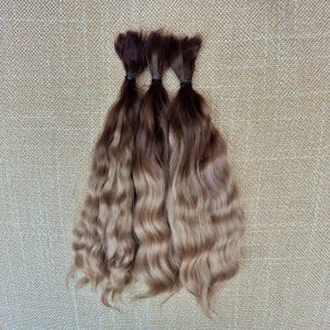 May include: Three bundles of wavy hair extensions featuring a dark brown root color that transitions into a lighter ash blonde shade at the ends. Each bundle is secured at the top with a small black elastic band, displaying a smooth texture.
