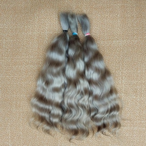 May include: Three bundles of wavy, gray hair extensions with dark bands at the top, each secured with a different color band: black, blue, and pink. The hair has a natural, wavy texture and is arranged on a textured, tan surface.