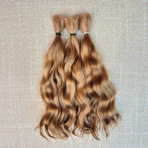 May include: Three bundles of wavy, light brown hair extensions. Each bundle is secured at the top with a small elastic band, featuring a natural gradient of golden and chestnut tones throughout the length of the strands, creating a soft texture.