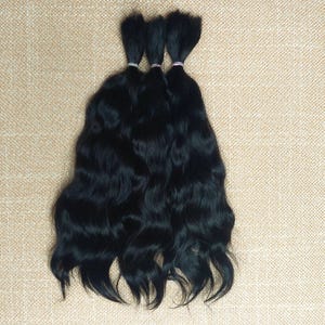 May include: Three bundles of wavy, black hair are displayed against a neutral background. Each bundle is tied at the top with a small, colorful elastic band. The hair appears to be of high quality, with a natural wave pattern.