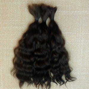 May include: Three bundles of dark brown wavy hair, each secured with a black elastic band. The hair has a natural, textured appearance, with visible waves and curls. The background is a neutral, textured surface.