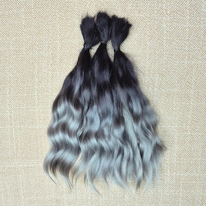 May include: Three bundles of wavy hair extensions featuring a dark black root color that transitions into a light silver-gray ombre effect. Each bundle is secured at the top with a small black band, showcasing a smooth texture at the roots.