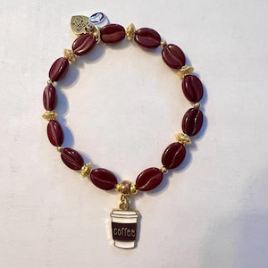 Könnte beinhalten: Ein burgunderfarbenes Perlenarmband mit goldenen Akzenten und einem Kaffeebecher-Anhänger. Das Armband hat kaffeebohnenförmige Perlen und kleine goldene Perlen. Der Anhänger sagt "coffee" auf einer weißen Tasse mit einem braunen Ärmel.