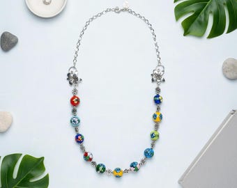 May include: A silver necklace featuring patterned glass beads in various colours. The necklace has a chain with flower-shaped clasps and small floral accents. The text "Ivy Lane Jewelry Shop" is at the bottom.