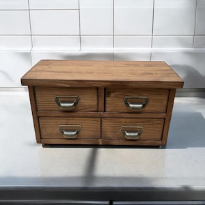May include: A small, wooden chest of drawers with four drawers. The chest is made of light brown wood and has antique-style metal handles. The drawers are arranged in two rows of two. The chest sits on a flat surface.
