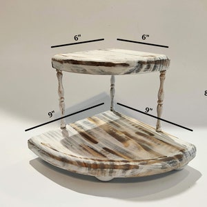May include: Whitewashed wooden corner shelf with two tiers. The top tier is 6 inches wide and the bottom tier is 9 inches wide. The shelf is 8 inches tall.