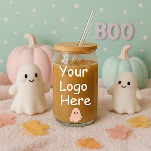 May include: A glass tumbler with a wooden lid and a clear straw, filled with a brown liquid. The text "Your Logo Here" is displayed on the front. The tumbler is surrounded by Halloween-themed decorations, including ghost figures and pumpkins.