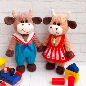May include: Two knitted brown and white toy cows, one wearing a blue sailor outfit and the other wearing a red and white dress with a bell and rhinestones.