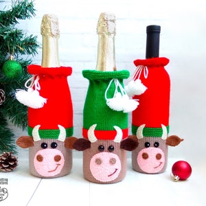 May include: Three wine bottle covers knitted in the shape of a cow. The covers are red, green, and brown with white pom-poms.