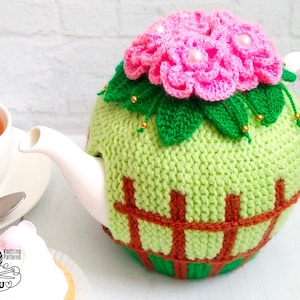 Puede incluir: Una funda de tetera de punto verde con un diseño de flores rosas y un patrón de valla marrón. La funda está en una tetera blanca.