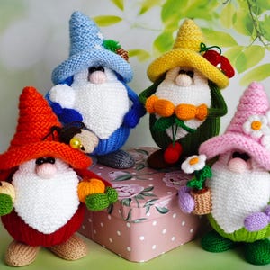 May include: Four knitted gnomes with white beards and colourful hats. One gnome wears a red hat and holds pumpkins, another has a blue hat, a third has a yellow hat with cherries, and the last has a pink hat with flowers. They are on a pink polka-dot surface.