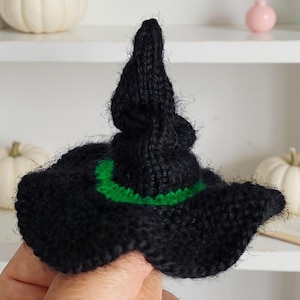 Può includere: Un cappello da strega nero lavorato a maglia con una fascia verde. Il cappello ha una parte superiore a punta e un'ampia tesa floscia. È un oggetto a tema Halloween o autunnale.