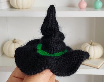 May include: A black knitted witch hat with a green band around the brim. The hat has a pointed top and a wide, floppy brim. The hat is a Halloween or fall-themed item.