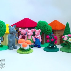 Puede incluir: Un conjunto de punto de tres personajes de cerdos, un personaje de lobo y un hongo, junto con una casa con techo rojo, una casa marrón, un árbol verde, un arbusto de bayas rojas y un arbusto de flores rosas. Los personajes y las casas están todos tejidos y tienen un aspecto caprichoso y hecho a mano.
