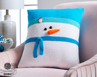 Può includere: Un cuscino a forma di pupazzo di neve in maglia bianca con un cappello, una sciarpa e strisce in maglia blu. Il pupazzo di neige ha occhi neri e un naso di carota arancione.