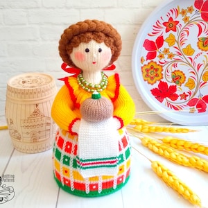 May include: A knitted doll wearing a yellow, red, green, and white dress with a white apron and holding a brown knitted pumpkin. The doll has brown hair and a red ribbon. The doll is sitting on a white surface with a wooden barrel and a tray with a floral design in the background.