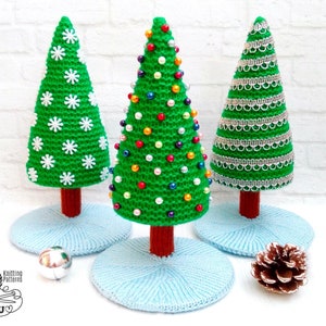 May include: Three crocheted Christmas trees, each decorated differently. One tree has white snowflakes, one has colorful beads, and one has silver trim. The trees are on blue crocheted bases.