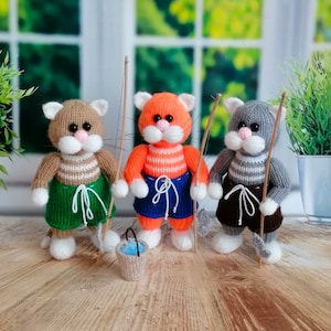 Puede incluir: Tres figuritas de gato de punto que llevan camisas a rayas y pantalones cortos, sosteniendo cañas de pescar y un cubo. Los gatos son marrones, naranjas y grises.