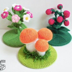 Puede incluir: Tres decoraciones de ganchillo: un ramo de flores rosas y blancas, un racimo de bayas rojas y tres setas naranjas y blancas. Cada decoración está sentada sobre una base de ganchillo verde.