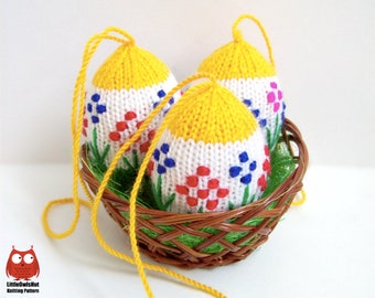 Può includere: Tre decorazioni di uova di Pasqua lavorate a maglia con top gialli, corpi bianchi e disegni floreali ricamati in rosso, blu e rosa. Le uova sono in un cestino di vimini marrone con un anello di corda gialla per appenderle.