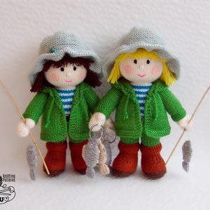 May include: Two knitted dolls wearing green jackets and hats, holding fishing rods and a string of fish. The dolls have brown and blonde hair, and are wearing brown boots.