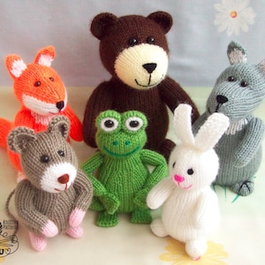 May include: A collection of six knitted animal toys: a brown bear, a green frog, a white rabbit, an orange fox, a grey mouse, and a grey wolf. The toys are all sitting on a white and green floral background.
