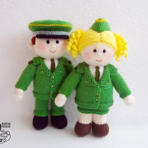 Puede incluir: Dos muñecas de punto que llevan uniformes verdes con botones dorados y sombreros. La muñeca femenina tiene el pelo rubio y la muñeca masculina tiene el pelo castaño.