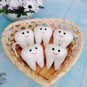 May include: Five white knitted tooth-shaped keychains with black eyes and red smiling mouths are displayed in a woven basket. The keychains are arranged in a circle with the basket in the center.
