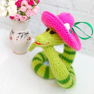 May include: A green and white striped knitted snake wearing a pink hat with a white flower accent. The snake is sitting on a white surface.