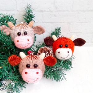 Puede incluir: Tres adornos de vaca tejidos en marrón y naranja con caras blancas y ojos negros. Los adornos están colgados de una rama de pino verde.