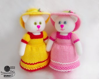Può includere: Due bambole di gatto a maglia che indossano abiti e cappelli. La gatta a sinistra indossa un vestito giallo con un bordo rosso e un cappello giallo. La gatta a destra indossa un vestito rosa con un bordo bianco e un cappello rosa.