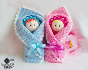 Può includere: Due bambole di lana avvolte in coperte, una blu e una rosa. Le bambole hanno capelli di lana e volti ricamati. Le coperte hanno bordi a uncinetto bianchi e sono legate con nastri abbinati.