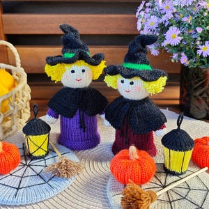 Può includere: Due bambole di strega fatte a mano con capelli gialli e cappelli, mantelli e vestiti neri. Le bambole sono circondate da zucche, lanterne e scope lavorate a maglia. Un cesto di zucchine gialle e un vaso di fiori viola sono sullo sfondo.