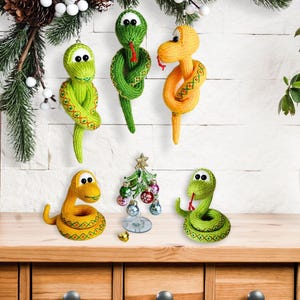 May include: A collection of handmade, knitted snake ornaments in shades of green and yellow. Some are hanging, others are coiled on a wooden surface. A small Christmas tree and decorative greenery are also present, suggesting a holiday theme.