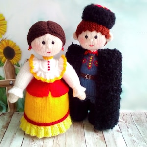 May include: Two knitted dolls wearing traditional Ukrainian clothing. The female doll is wearing a white blouse with a red and yellow skirt. The male doll is wearing a blue shirt with a black coat and a black hat.
