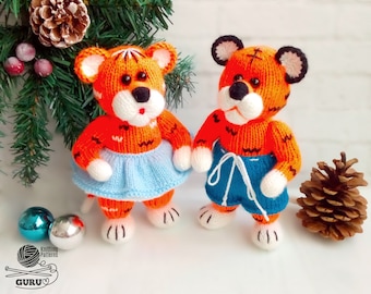 Può includere: Due peluche di tigre a maglia, uno con una gonna blu e l'altro con pantaloncini blu. Entrambi i peluche hanno strisce arancioni e nere e volti bianchi.