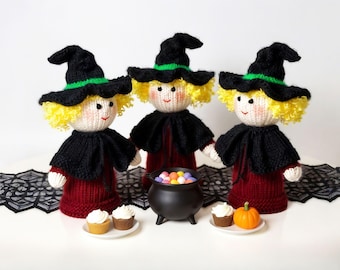 Può includere: Tre bambole di strega lavorate a maglia con cappelli neri, mantelli e vestiti rossi. Ogni bambola ha i capelli gialli e un viso sorridente. Un calderone nero con caramelle colorate, cupcakes e una piccola zucca. Decorazioni di Halloween.