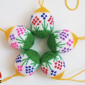 May include: Six knitted Easter egg decorations. Each egg has a white base with floral patterns in red, blue, and pink. The bottom is green, and the top is yellow, with a yellow hanging string. The eggs are handmade.