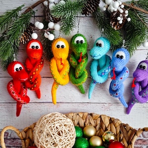 Pode incluir: Sete ornamentos de cobra de malha coloridos pendurados em um galho. As cobras são vermelhas, amarelas, verdes, azuis, azul claro, roxas e rosas. Todas estão enroladas em uma pose semelhante.