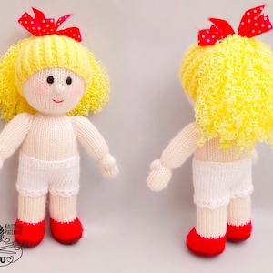 Peut inclure: Une poupée en tricot avec des cheveux bouclés jaunes, un nœud rouge et des vêtements blancs. La poupée est debout sur une surface blanche.
