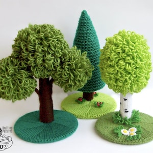 May include: Three knitted trees with green foliage and brown trunks. The trees are standing on green bases. The tree on the right has a white trunk with black markings and white flowers at the base.