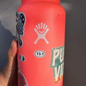 May include: A bright pink reusable water bottle with a white Hydro Flask logo and various stickers, including a sticker with the text "13.1" and a sticker with the text "PU" and "VI".