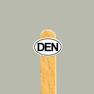 May include: A small oval sticker with a white background and a thin black border featuring the black text DEN in capital letters. The sticker is attached to the top portion of a light brown wooden craft stick with a rounded top edge.