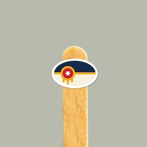 Op de afbeelding: Een ovale sticker met een horizontaal ontwerp met een donkerblauw bovengedeelte, een dunne gouden middenstreep en een crèmekleurig ondergedeelte. Een rood cirkelvormig embleem met een witte ster en drie naar beneden wijzende gouden druppels.
