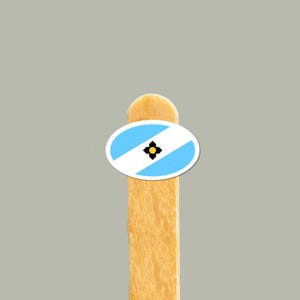 Op de afbeelding: Een ovale sticker met een diagonaal lichtblauw en wit gestreept patroon met een centraal zwart vierbladig bloempictogram met een geel hart, bevestigd aan een lichtbruin houten stokje.