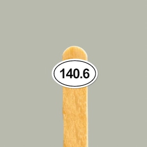 May include: A wooden popsicle stick with a white oval sticker displaying the number 140.6 in black. The stick is light brown, and the background is a solid gray.