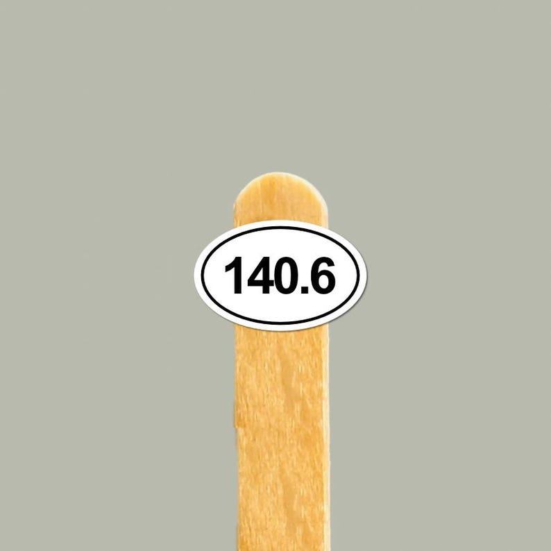 May include: A wooden popsicle stick with a white oval sticker displaying the number 140.6 in black. The stick is light brown, and the background is a solid gray.