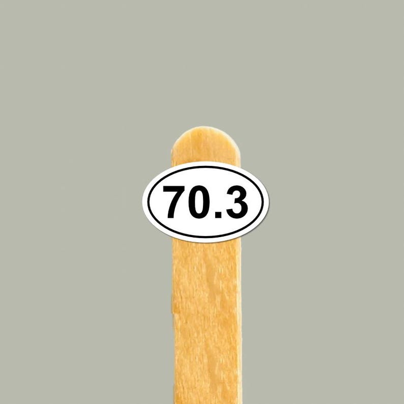 May include: A wooden popsicle stick with a white oval sticker displaying the number "70.3" in black. The background is a solid, light gray color.
